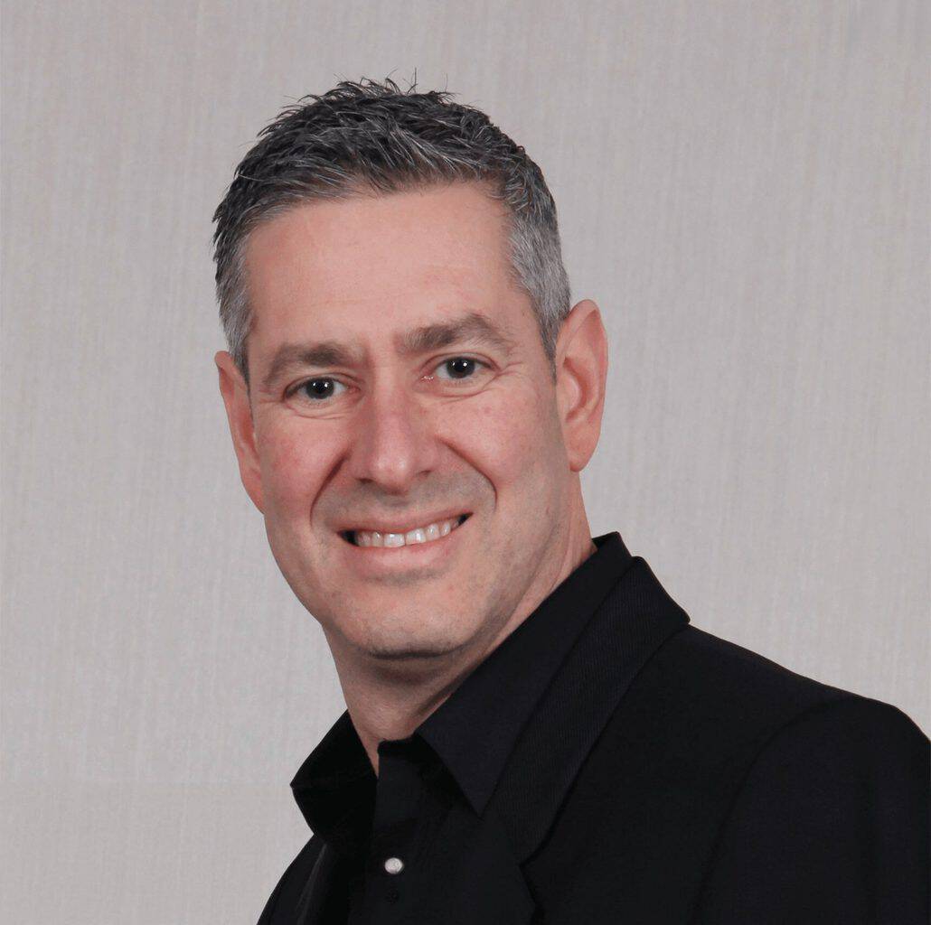 Close-up of a smiling man with short, dark hair wearing a black suit and shirt, against a neutral background, suitable for business or professional context.