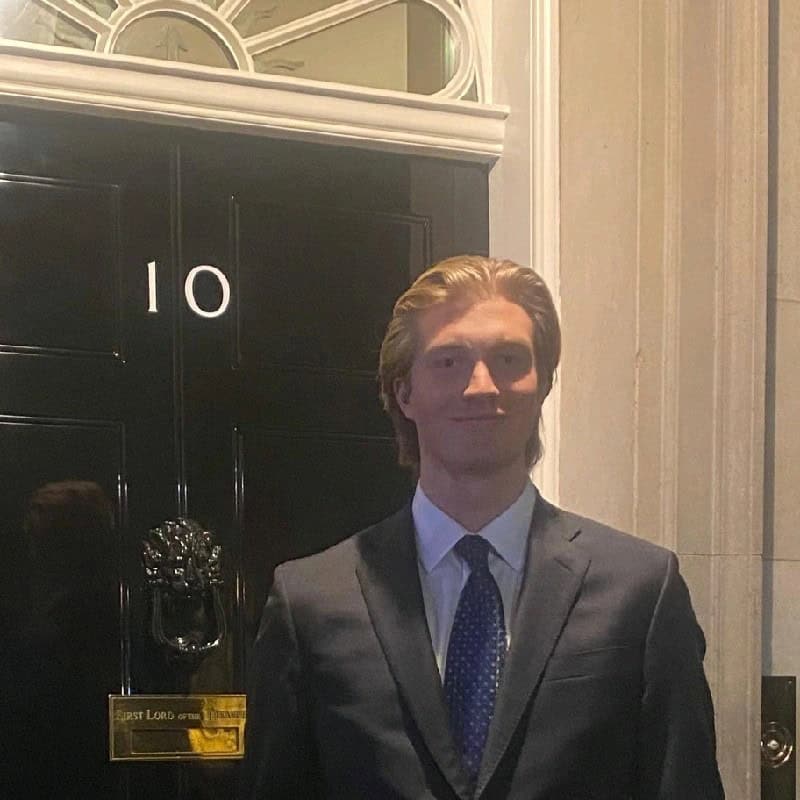 Young man in a formal suit standing outside a building with a black door and lion head door knocker, smiling at the camera.