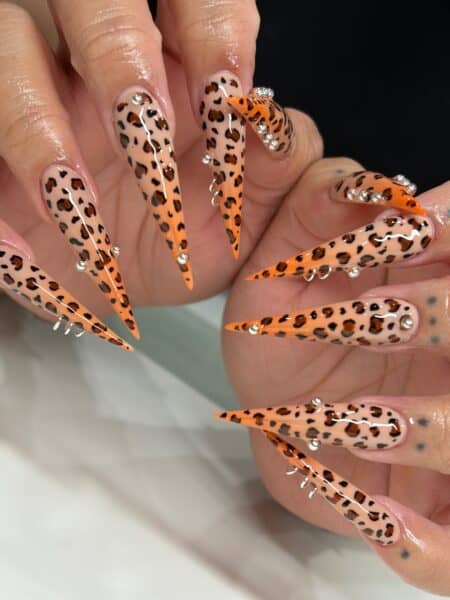 Leopard-print stiletto nails with orange tips and rhinestones