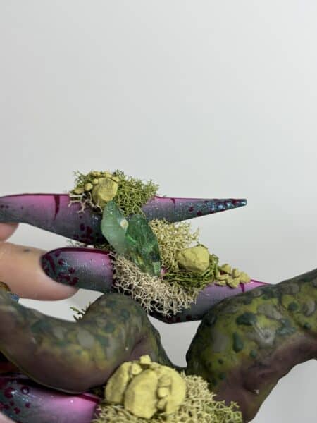 Glittery purple and green nails adorned with dried moss