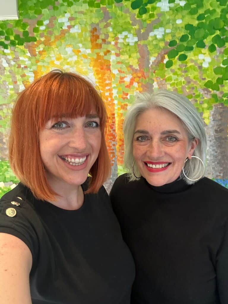 Two smiling women pose before a colourful abstract painting.