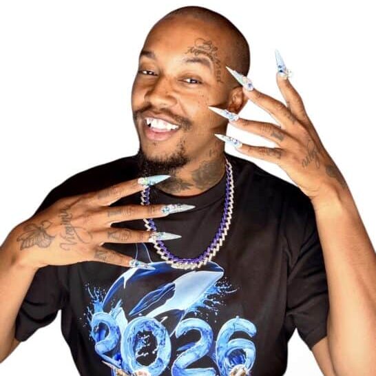 Calvin Jones showcasing elaborate nail art and tattoos, smiling confidently, wearing a black t-shirt with '2026' printed on it, and a purple beaded necklace.