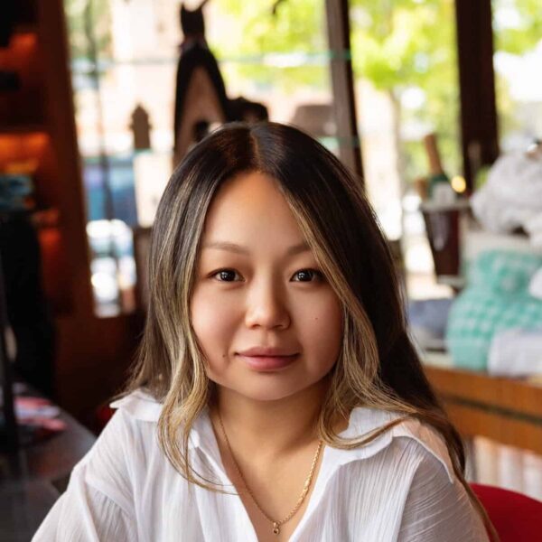 Young woman with long dark hair and a white blouse sitting at a table in a cafe, with a coffee cup in front of her, smiling softly at the camera.