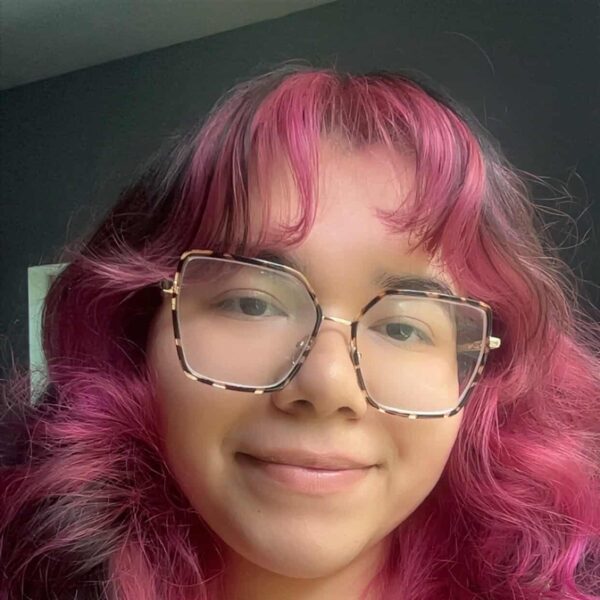 Young woman with pink curly hair, glasses, and a grey top smiling at the camera, showcasing vibrant hair color and style.